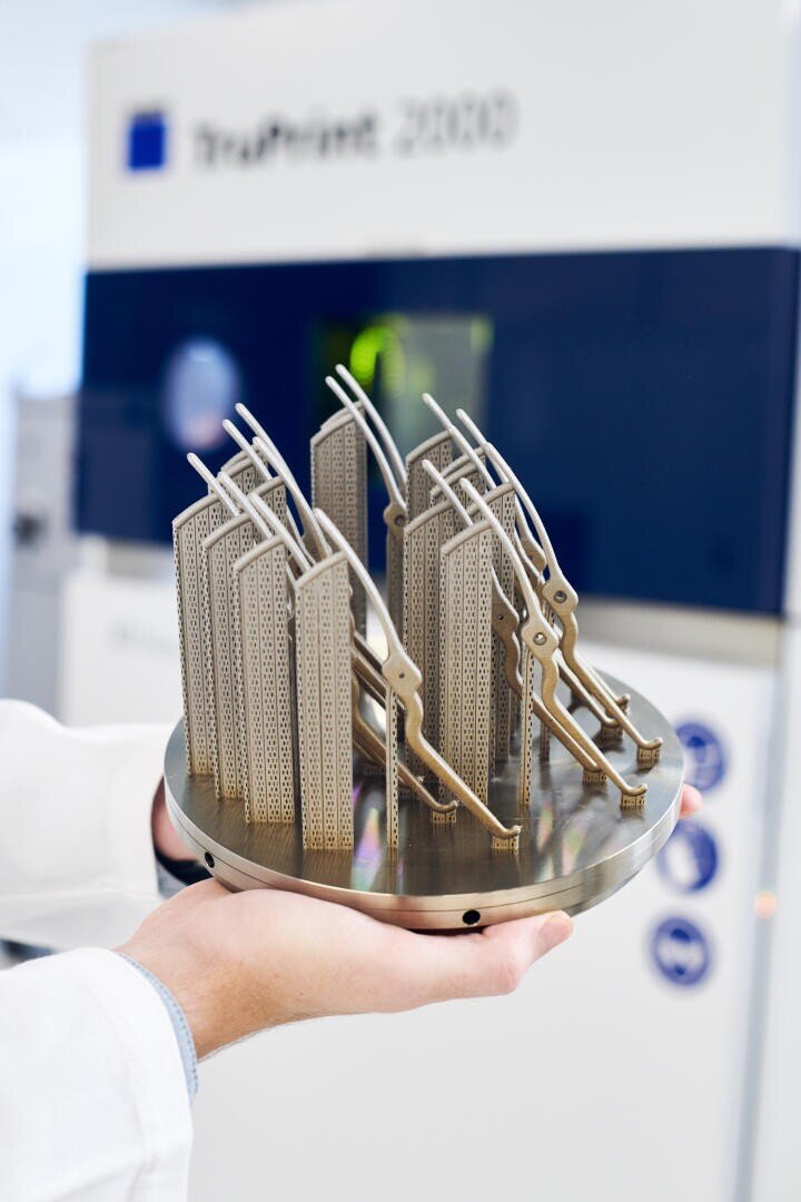 A person in a white lab coat holds a metal plate with intricate, lattice-like 3D-printed metal parts in front of a TruPrint 2000 3D printer in a laboratory setting.