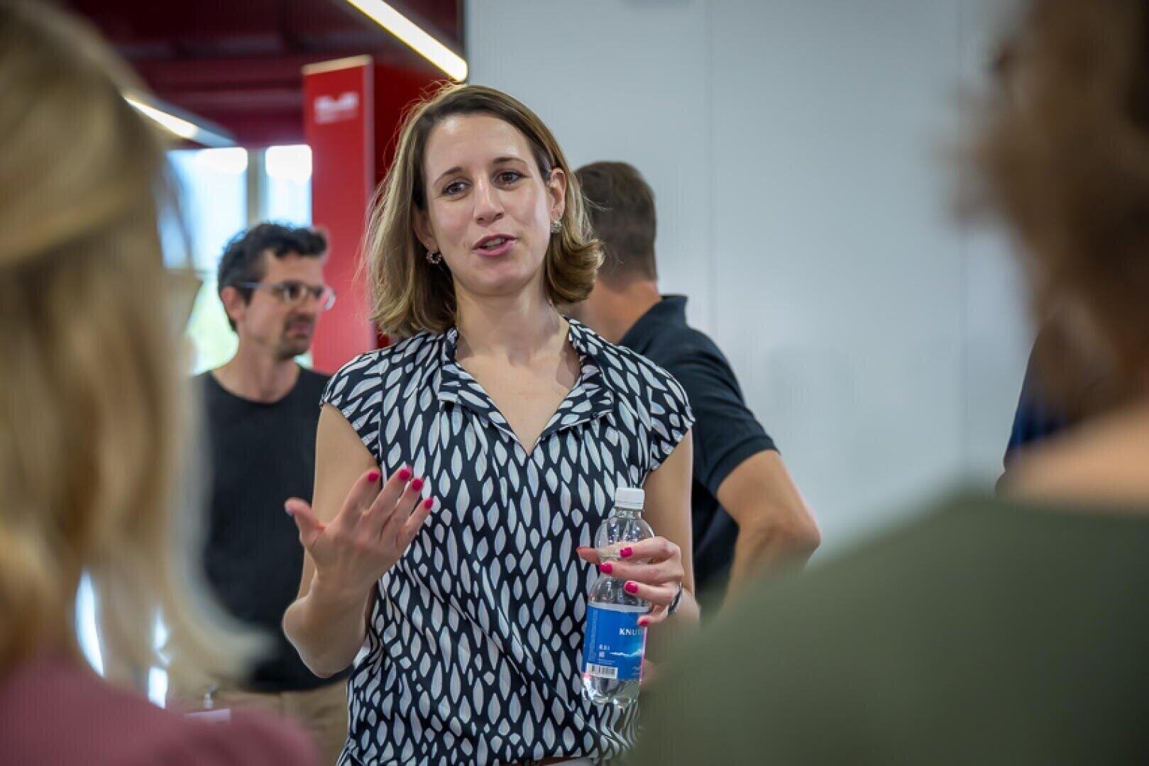 A woman holding a water bottle speaks to two people in the foreground. She gestures with her hand, appearing engaged in conversation. Several other people are visible in the blurred background of a brightly lit indoor space.