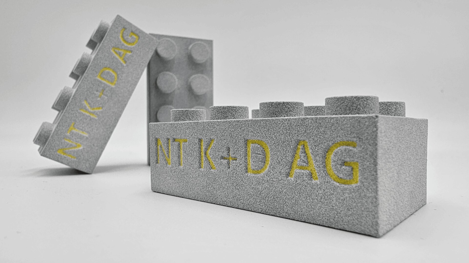 Three gray LEGO-style bricks with a rough texture stand on a white background. The front brick displays the yellow text NT K-DAG while the others are stacked and positioned in the background.