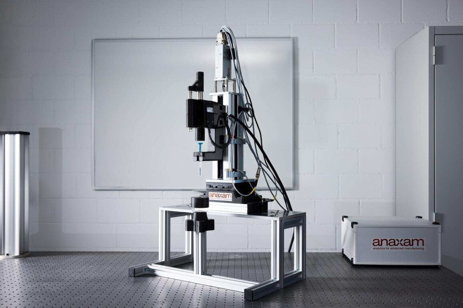 Eine Präzisions-Labormaschine mit Kabeln und einem Roboterarm steht auf einem Metallrahmen in einem sauberen, weißen Raum mit einer leeren Tafel und einer Kiste mit der Aufschrift Anaxam im Hintergrund.