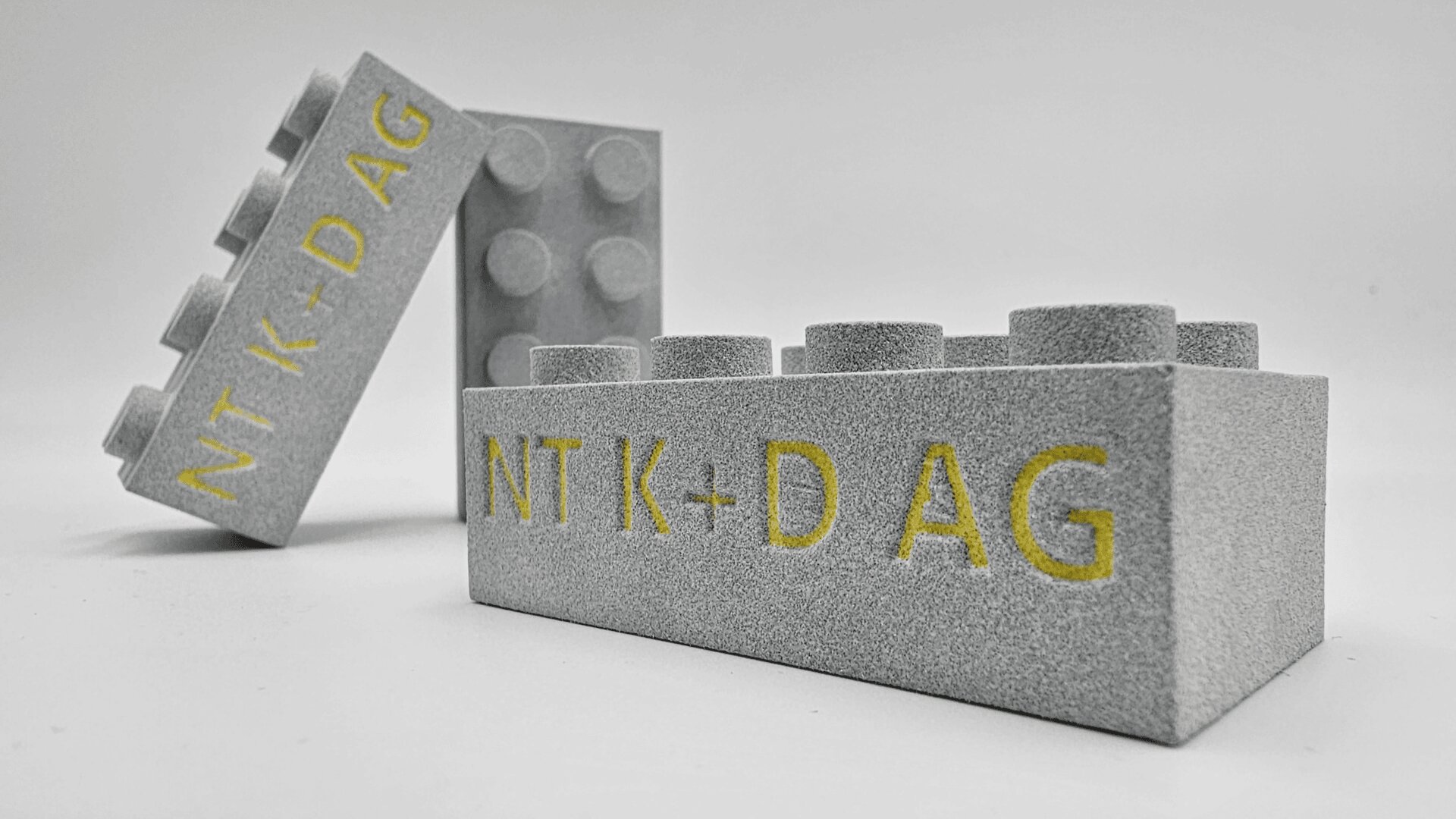 Drei graue Lego-ähnliche Blöcke mit rauer Textur sind auf weißem Untergrund angeordnet. Der vordere Block trägt den gelb eingravierten Text NT K-D AG. Die anderen Blöcke sind dahinter und seitlich positioniert.