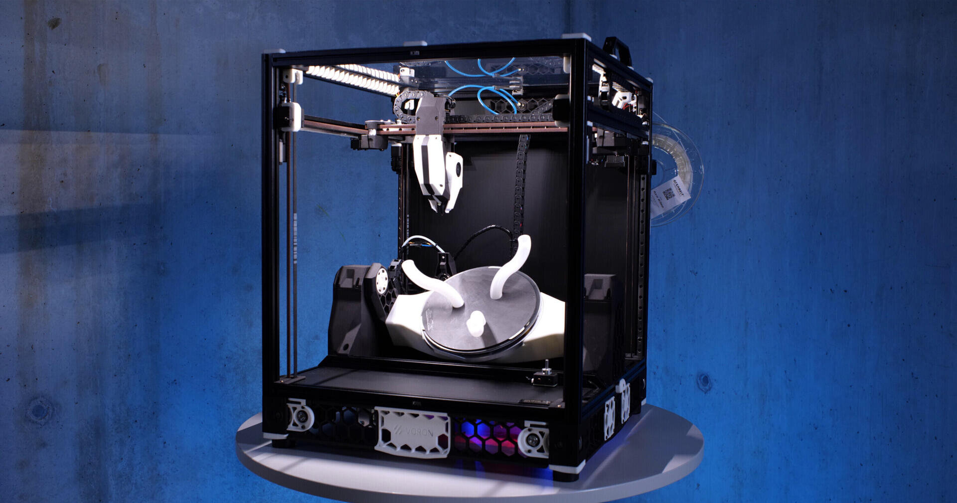 Ein 3D-Drucker steht auf einem runden Tisch und druckt ein weißes, gebogenes Objekt. Der Hintergrund ist eine blaue Wand, und im offenen Rahmen des Druckers sind verschiedene Drähte und Komponenten zu sehen.