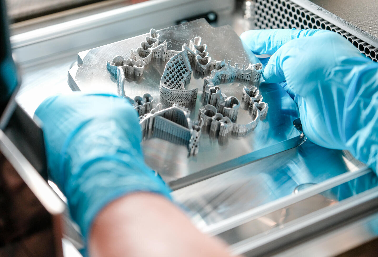 Mit Handschuhen wird ein metallisches 3D-gedrucktes Objekt mit verschlungenen, architektonischen Formen auf einer Metallplattform in einer modernen Fertigungs- oder Laborumgebung angefasst.