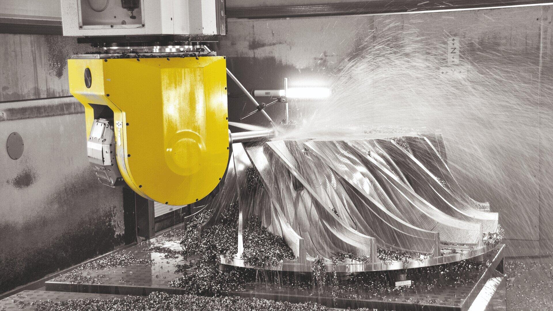 A CNC milling machine with a yellow head machines a large, complex metal part, spraying coolant onto the workpiece. The metal chips are scattered on the table.