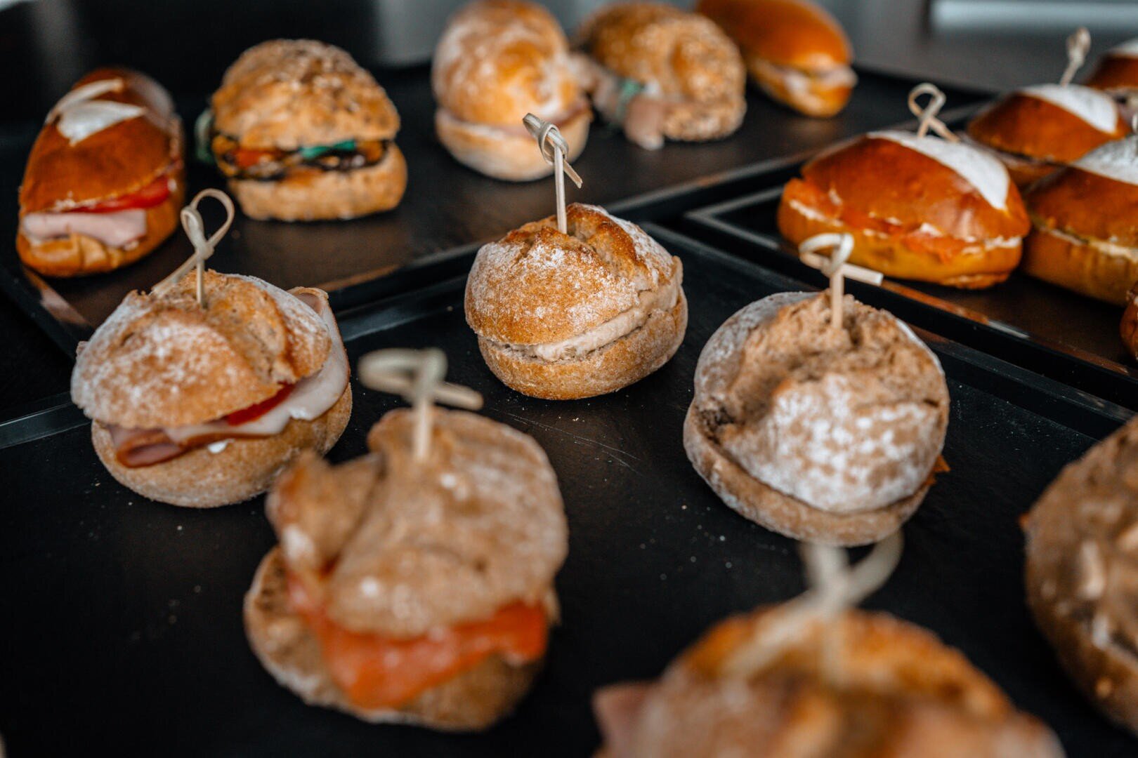 Auf schwarzen Tabletts sind verschiedene Sandwiches auf Vollkorn- und Laugenbrötchen angeordnet, die jeweils mit einem dekorativen Zahnstocher befestigt sind. Einige sind mit Mehl bestäubt und mit verschiedenen Fleischsorten und Grünzeug gefüllt.