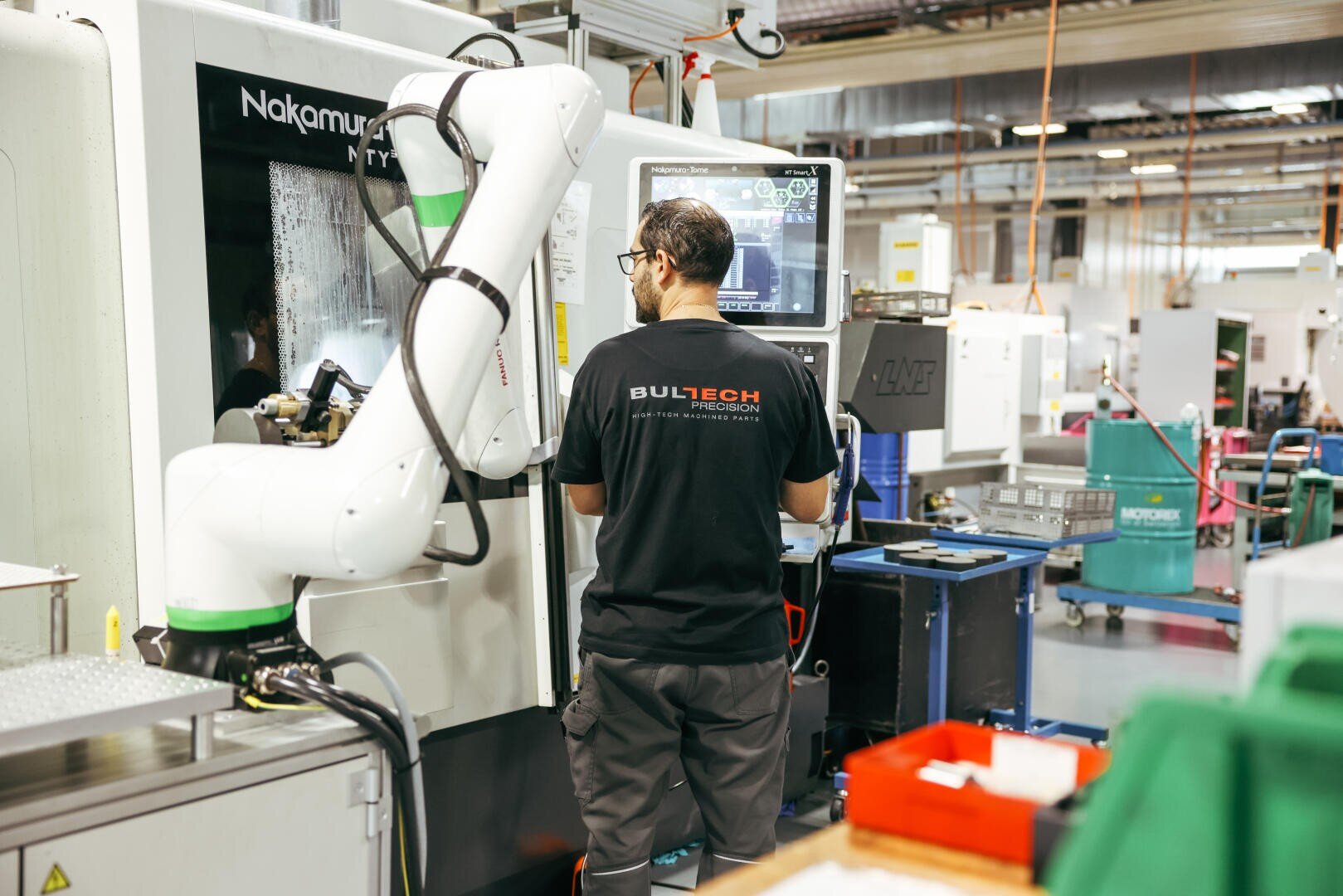 A person stands in front of a large machine and a monitor in a factory and operates a device with a visible robotic arm. The worker is wearing a black "BULTECH" shirt and safety goggles, surrounded by industrial tools.