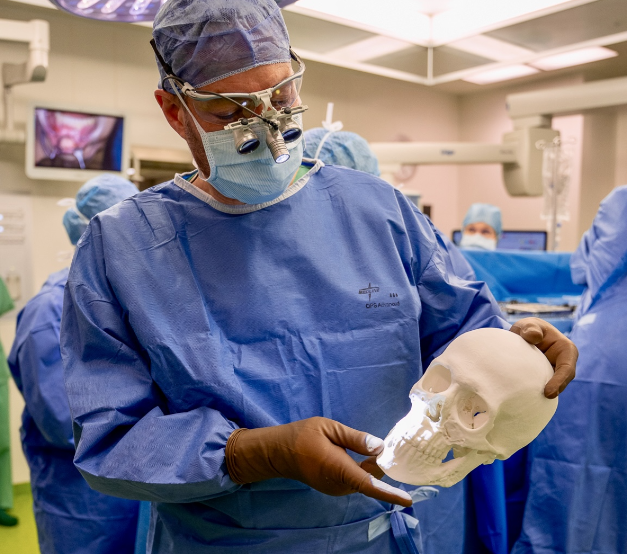 A surgeon in a blue coat and gloves holds a white 3D-printed model of a human skull in an operating theater, while medical staff and equipment can be seen in the background.