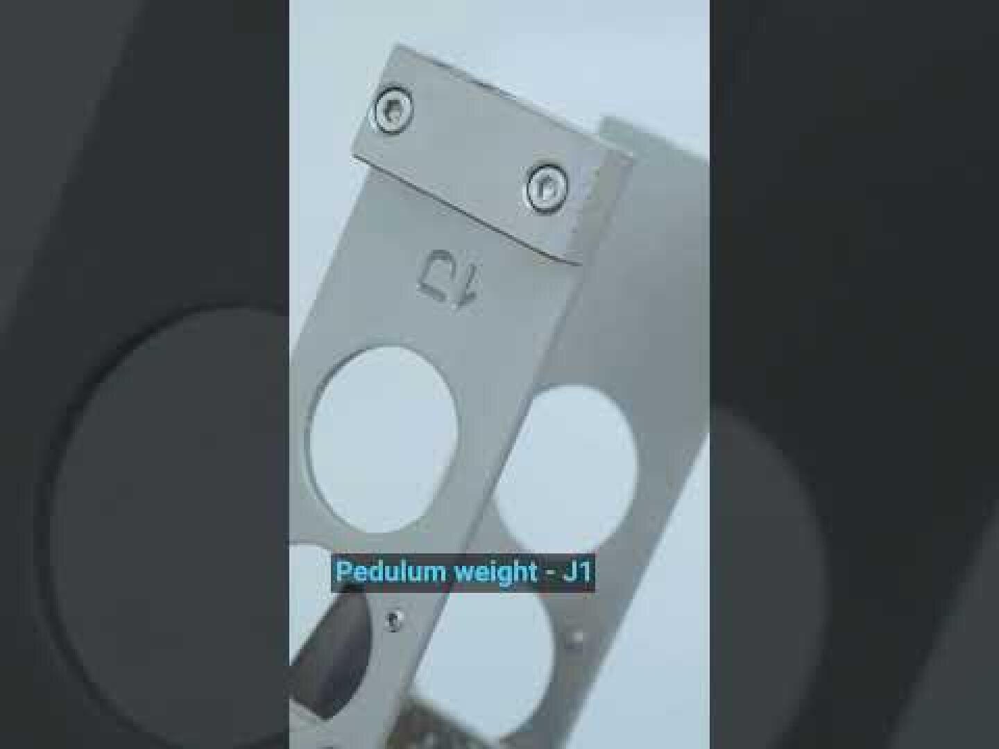 Nahaufnahme eines metallischen Pendelgewichts mit der Bezeichnung J1, mit kreisförmigen Ausschnitten und zwei sichtbaren Schrauben an der Oberseite. Die blaue Beschriftung lautet "Pedulum weight - J1".