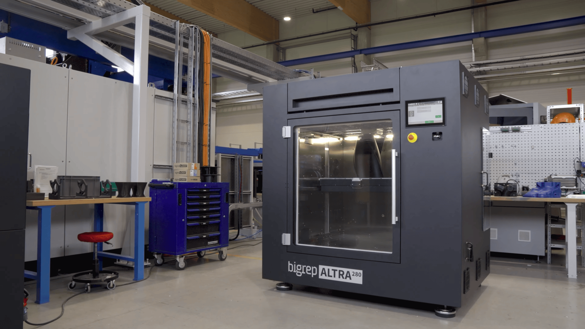 Ein großer schwarzer 3D-Drucker mit der Aufschrift bigrep ALTRA 280 steht in einer Industriehalle, umgeben von Werkbänken, Schränken und verschiedenen Werkzeugen und Geräten.