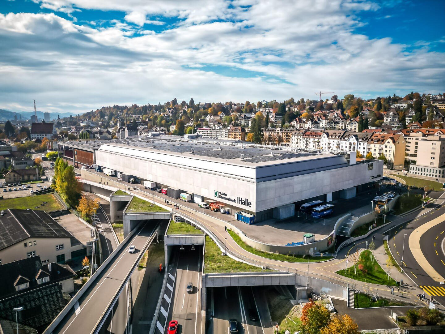 Ein großes Industriegebäude, vor dessen Laderampen Lastwagen geparkt sind, umgeben von Straßen und Überführungen, im Hintergrund ein Wohnviertel und Bäume unter einem teilweise bewölkten Himmel.