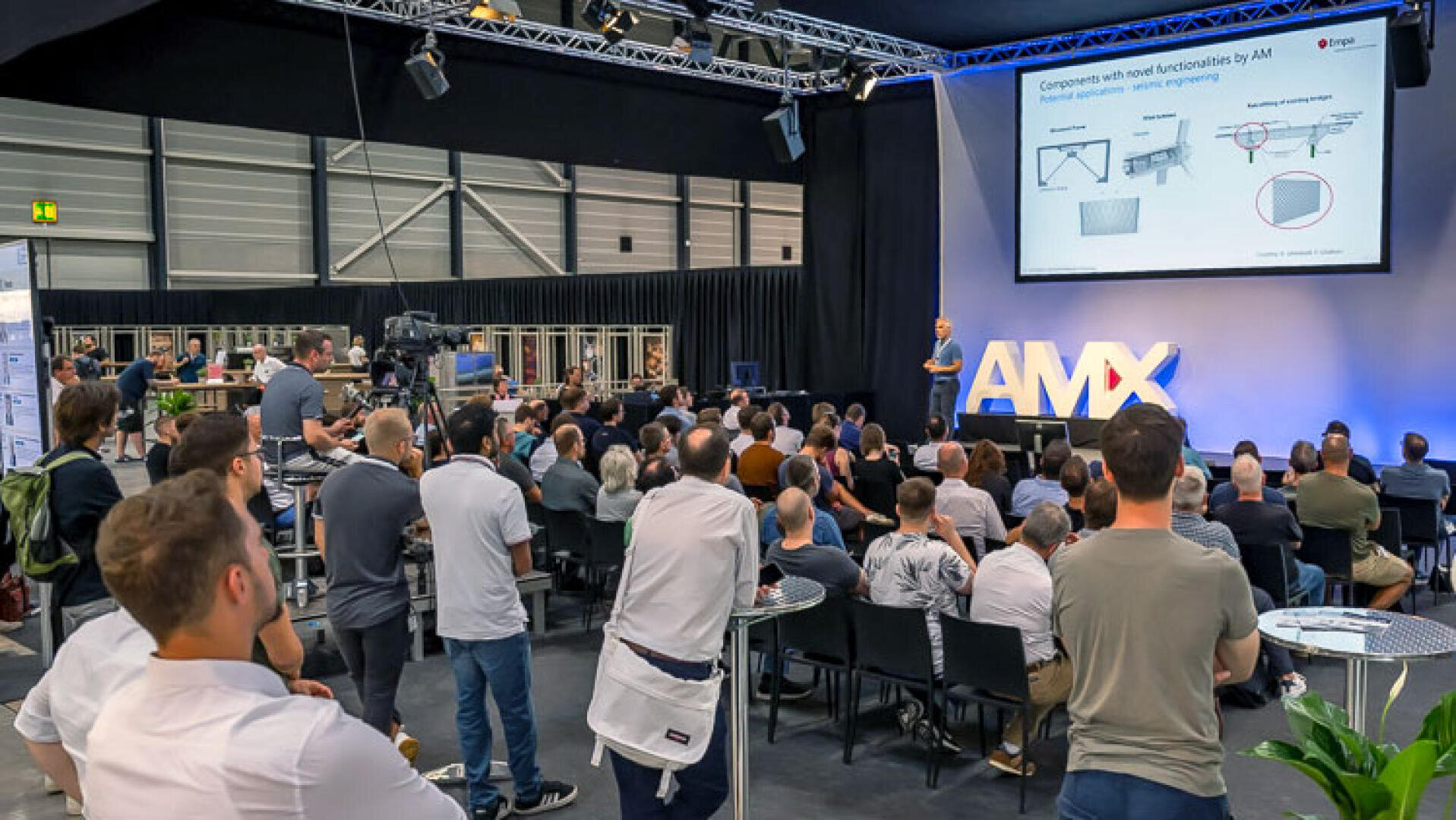 Ein Redner präsentiert einem sitzenden Publikum auf einer Konferenz eine technische Diashow. Auf der Bühne stehen große AMX-Buchstaben, und die Teilnehmer schauen in einem geräumigen, modernen Veranstaltungsort aufmerksam zu.