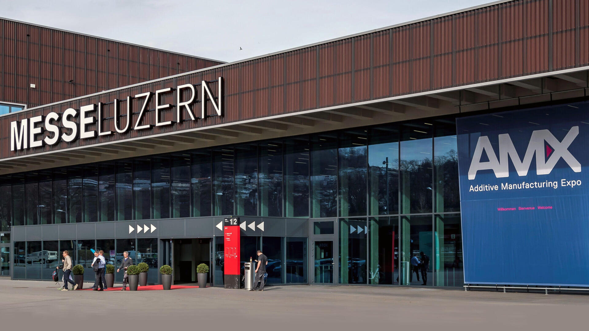 Large modern building with the sign MESSE LUZERN above the entrance. Several people are standing or walking outside. A large blue banner on the right-hand side advertises the AMX Additive Manufacturing Expo.