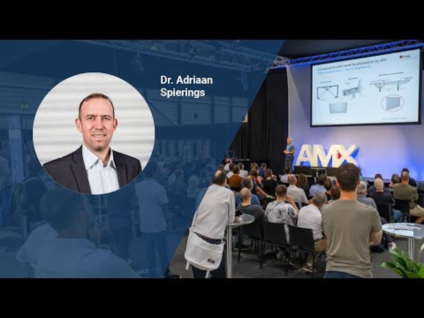 A professional event with an audience seated facing a stage where a speaker presents slides on a screen. An inset shows a portrait of Dr. Adriaan Spierings with his name beside it.