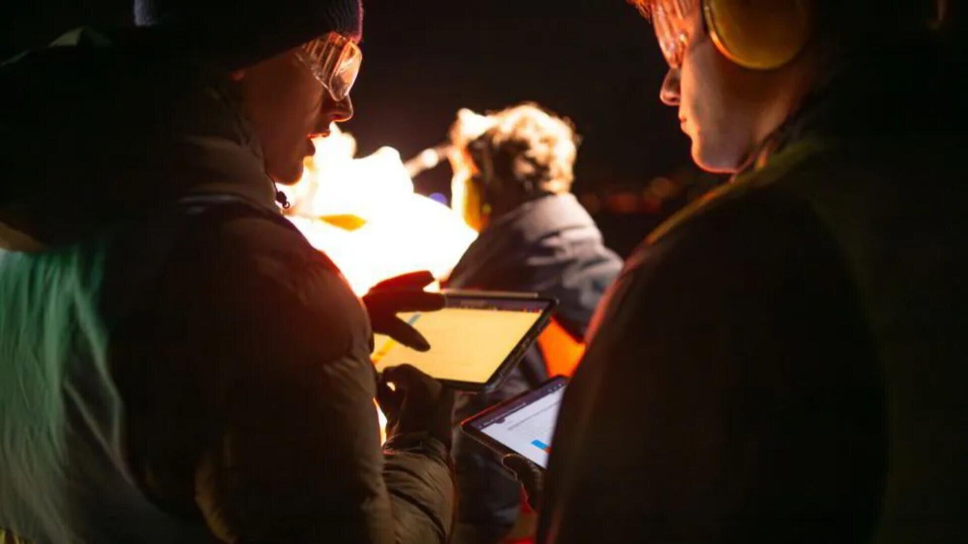 Zwei Personen mit Schutzkleidung und Brille stehen bei schwachem Licht und verwenden Tablets. Eine Person gestikuliert in Richtung ihres Tablets, während die andere zuhört und auf ihren eigenen Bildschirm schaut. Ein hell erleuchteter Hintergrund hebt eine weitere Figur hervor.