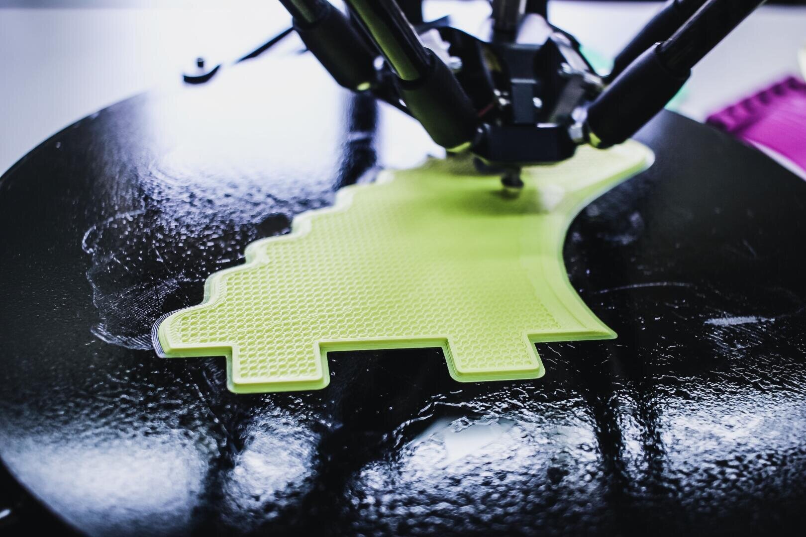 Eine Nahaufnahme eines 3D-Druckers, der ein leuchtend gelbes Objekt auf einer schwarzen Plattform erstellt. Das Objekt hat eine gezackte, verpixelte Form, und die mechanischen Arme des Druckers sind darüber sichtbar.