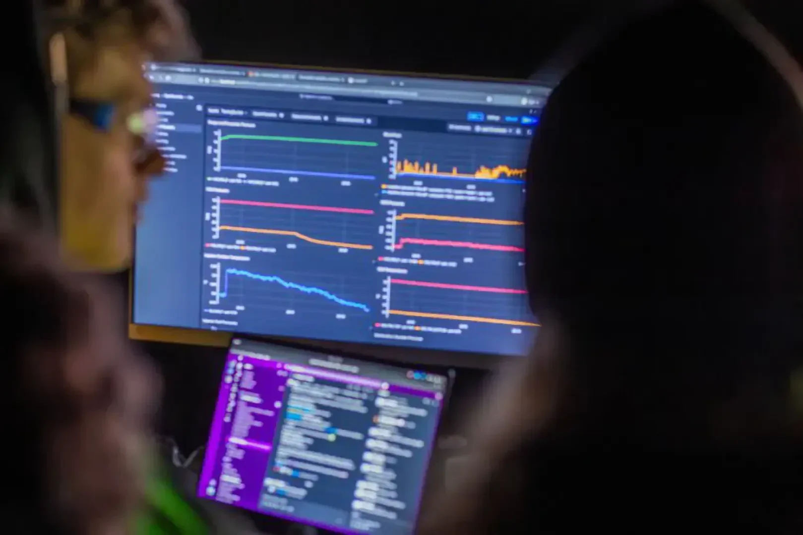 Zwei Personen blicken auf einen Computermonitor, auf dem mehrere farbige Liniendiagramme und Messwerte angezeigt werden. Darunter befindet sich ein Laptop, auf dem Codezeilen zu sehen sind, was auf eine Datenanalyse- oder Programmierumgebung schließen lässt.