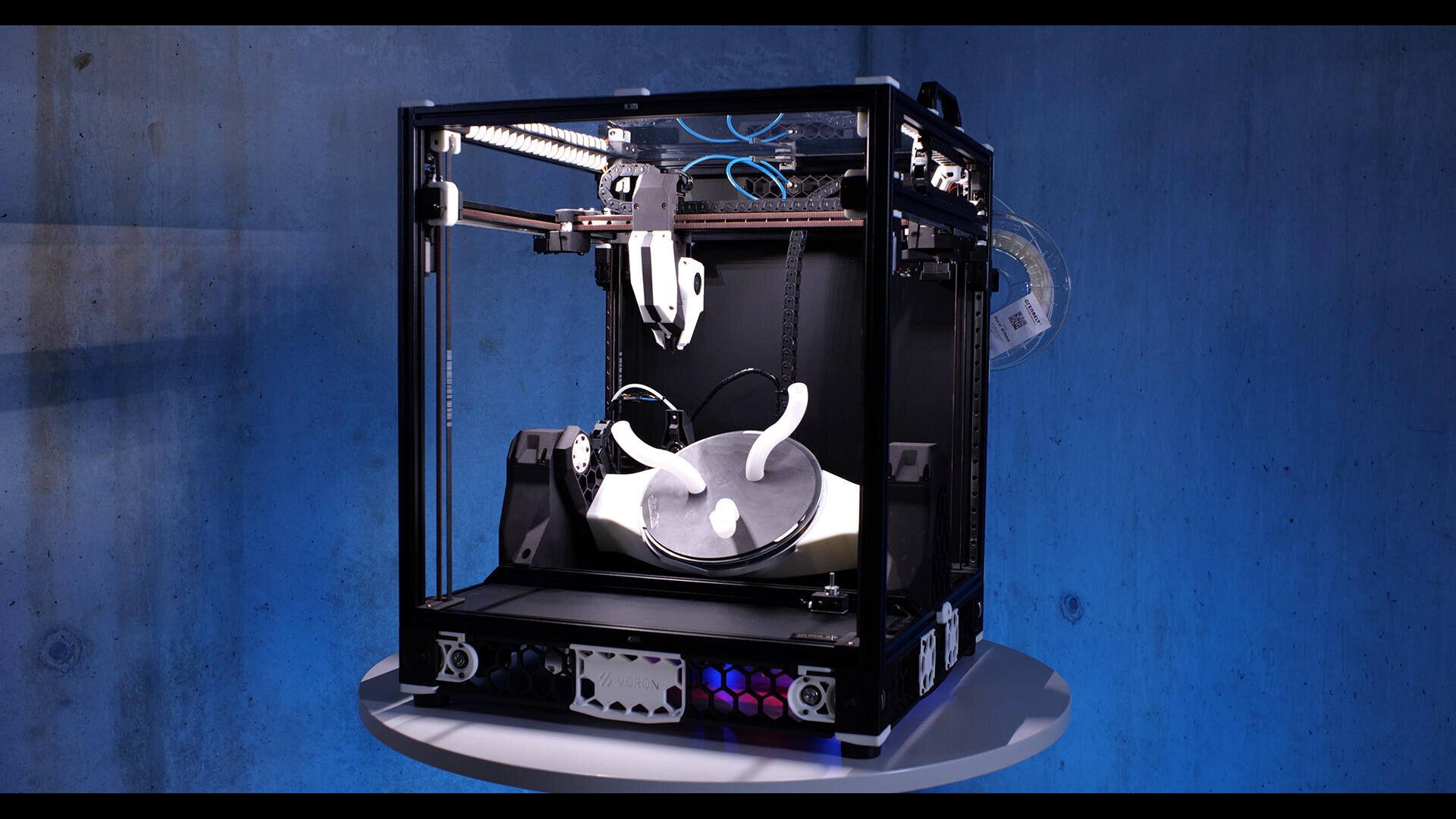 Ein 3D-Drucker auf einem runden Tisch druckt ein großes weißes Objekt mit einer geschwungenen, abstrakten Form. Der Hintergrund ist blau mit einer strukturierten Wand. Der Drucker hat einen schwarzen Rahmen und sichtbare Kabel.