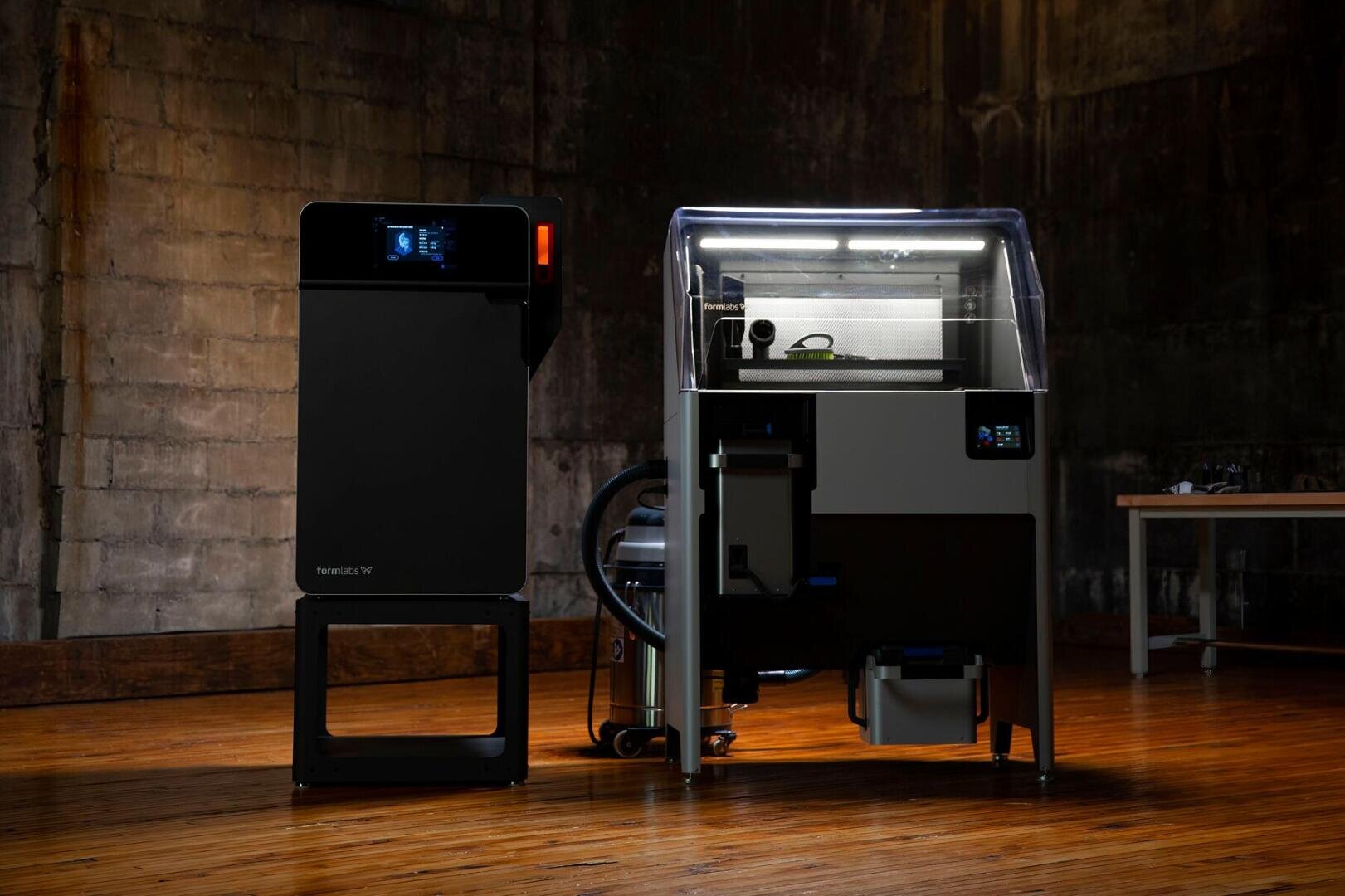 Two black 3D printing machines stand on a wooden floor in a dimly lit industrial space with concrete walls. One machine has a transparent cover and visible internal parts, the other has a digital display.