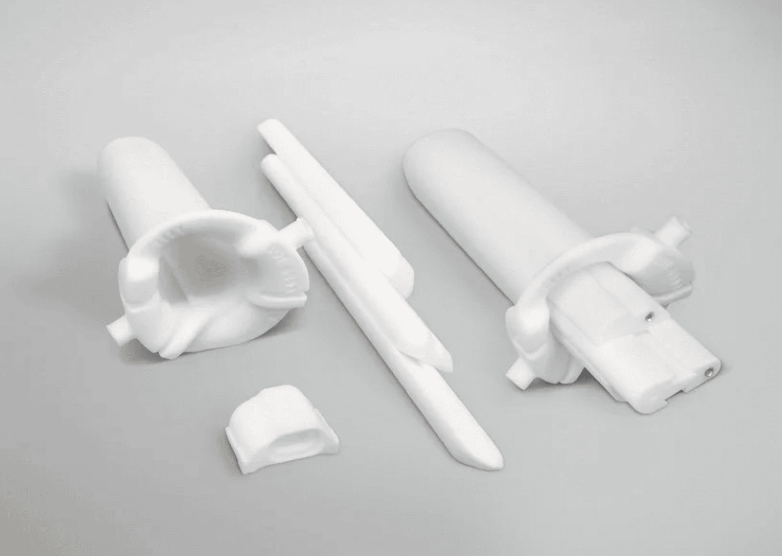 Five white plastic parts of varying shapes and sizes are arranged on a light gray background. The pieces include cylindrical components, flat elongated rods, and a small block-like piece.