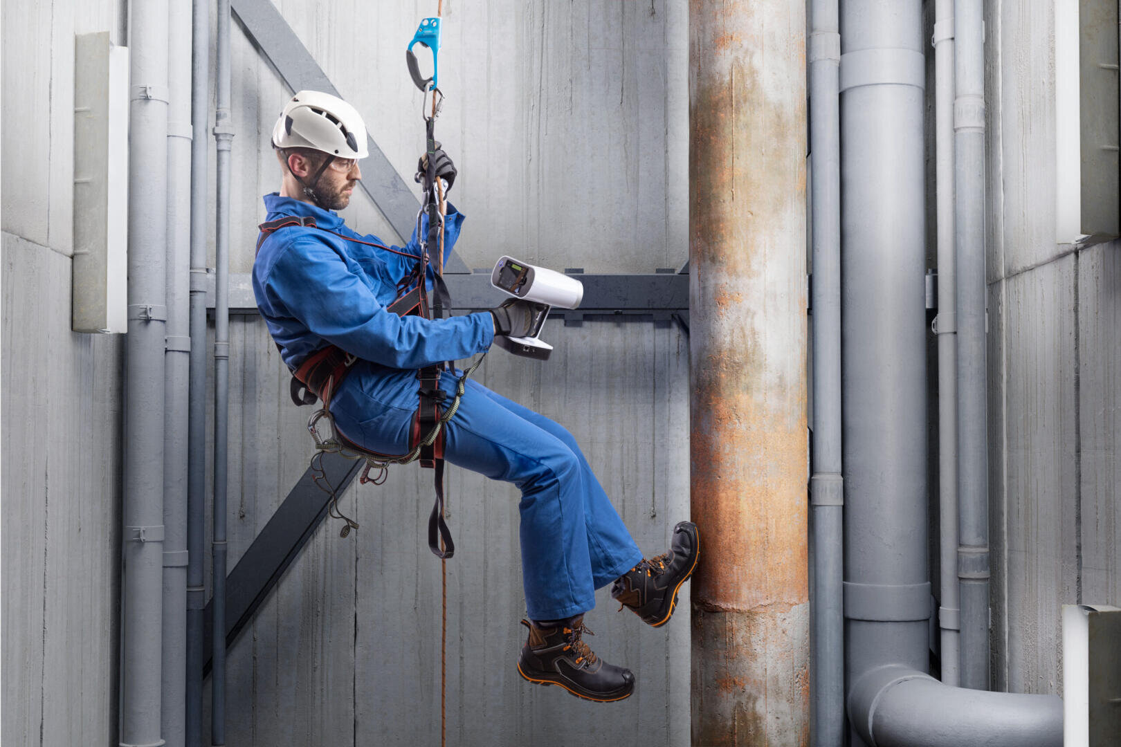 Ein Arbeiter in blauem Overall und Sicherheitsausrüstung ist an einem Gurt aufgehängt und inspiziert Rohre und Haltevorrichtungen in einer industriellen Umgebung.