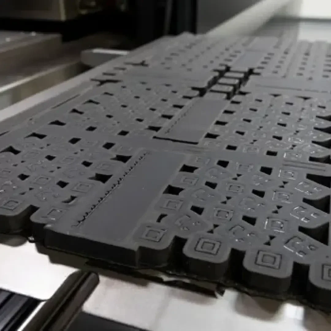 Nahaufnahme einer dunklen Gummimatte mit erhabenen quadratischen und rechteckigen Mustern auf einer metallischen Oberfläche, wahrscheinlich Teil eines industriellen Herstellungs- oder Druckverfahrens.