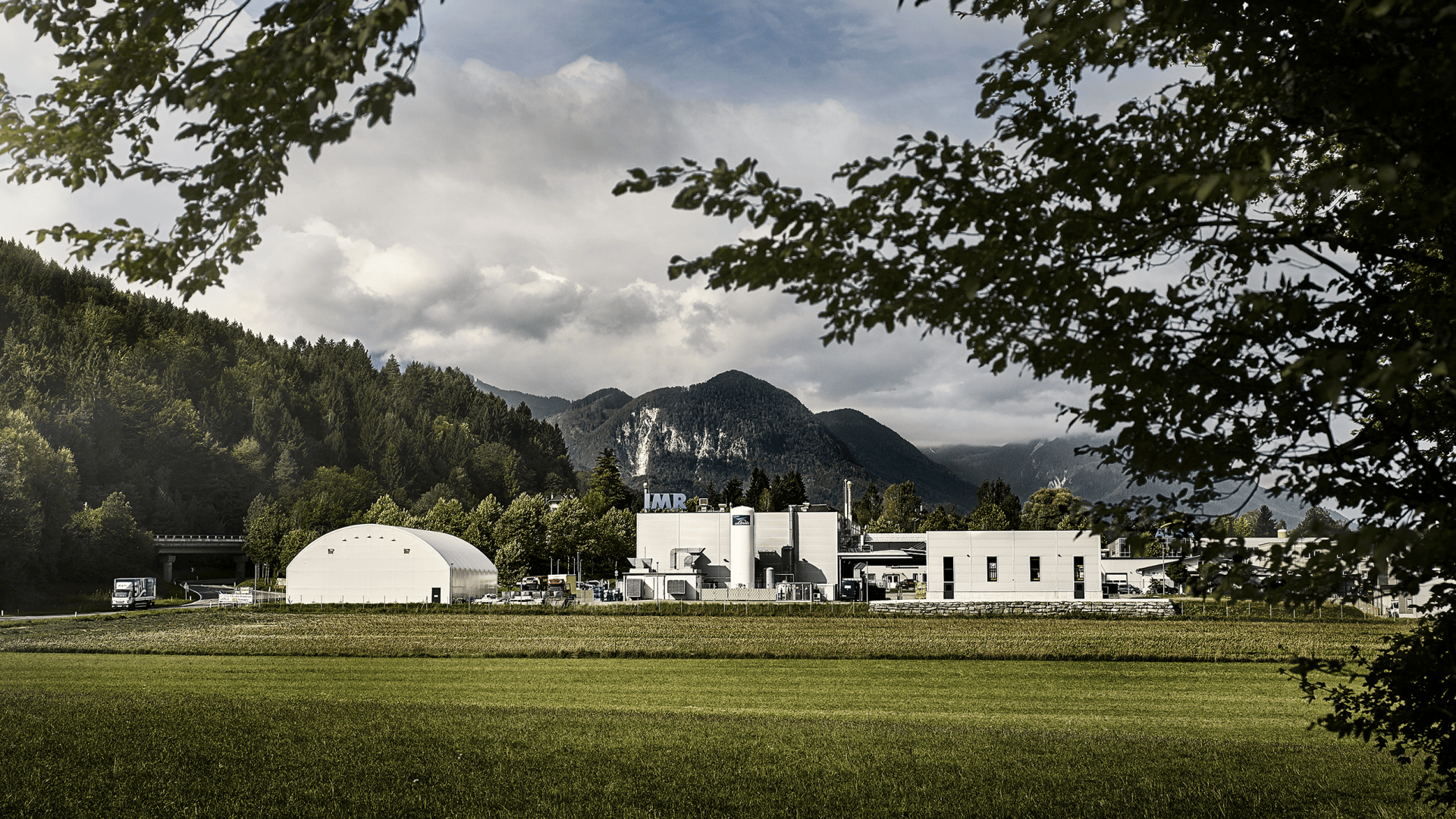 Eine moderne Industrieanlage mit weißen Gebäuden und Silos liegt vor einer Kulisse aus bewaldeten Hügeln und Bergen unter einem wolkenverhangenen Himmel, eingerahmt von grünen Bäumen und einer offenen Wiese.