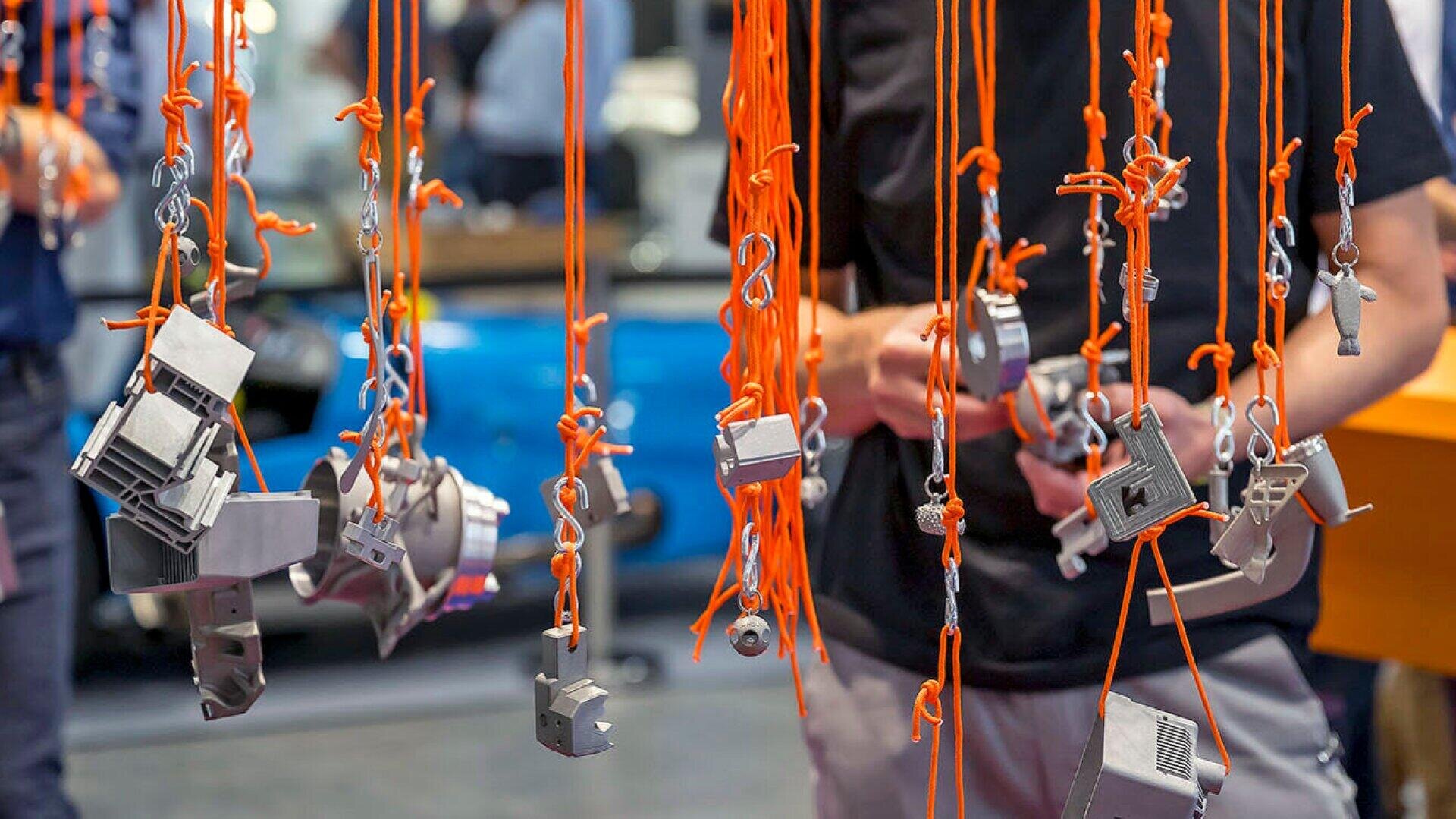 Numerous small metal objects hang from bright orange strings, which are suspended in front of people standing in a room. The scene appears to be at an exhibition or event, with a blue object blurred in the background.