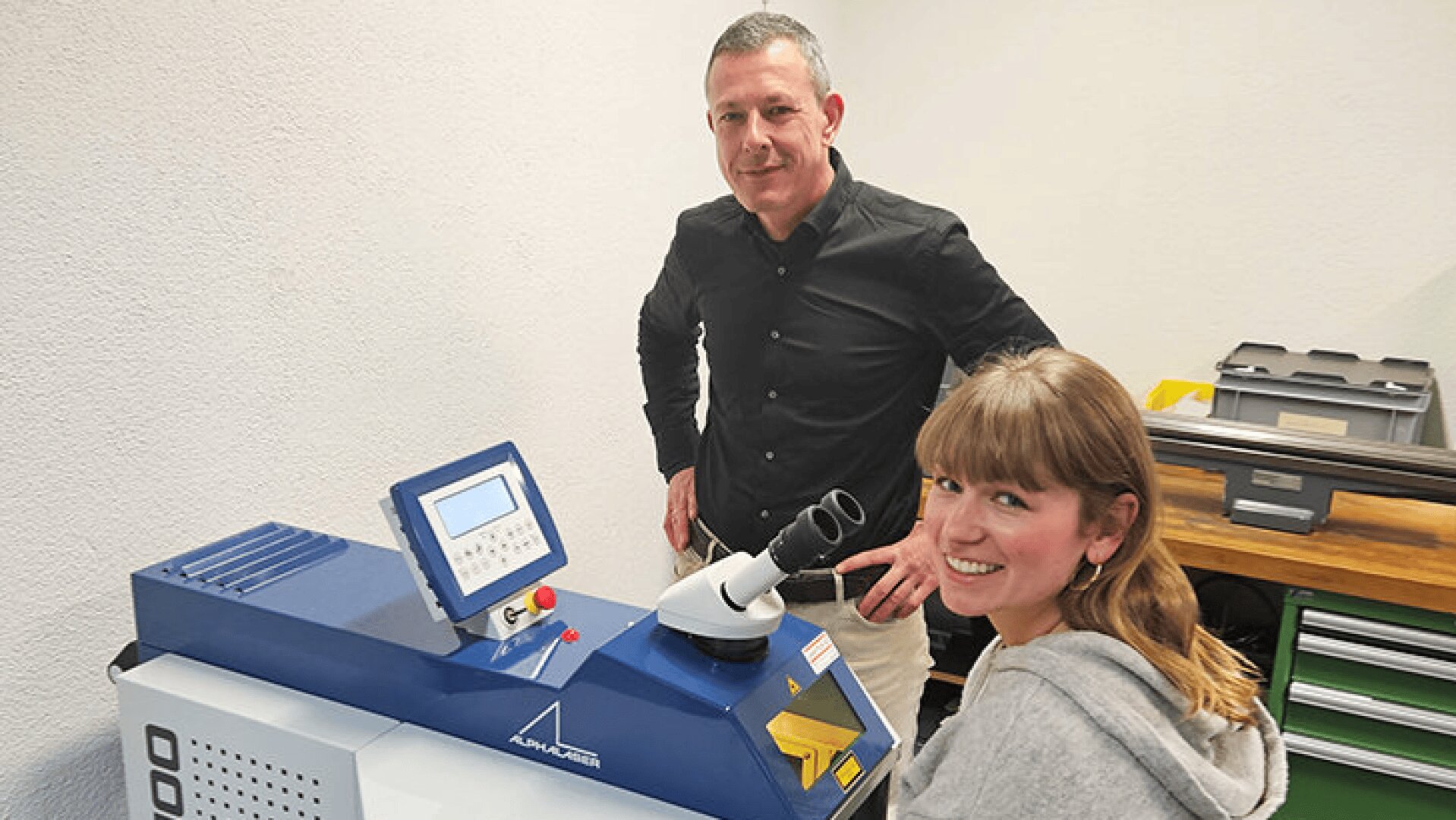 Ein Mann steht neben einer lächelnden Frau, die an einem blau-weißen Laborgerät mit digitaler Anzeige und Mikroskopaufsätzen in einem Arbeitsbereich sitzt und arbeitet.