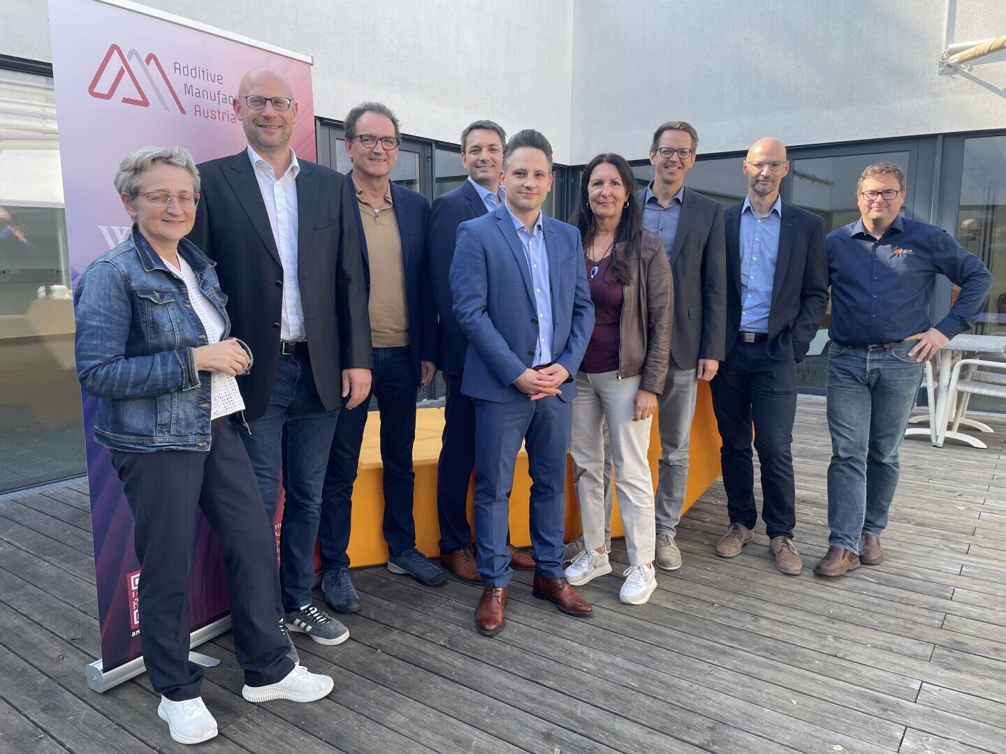 Nine people in business attire stand together on a wooden deck and smile into the camera. A banner with the words Additive Manufacturing Austria can be seen on the left. The group is outdoors with sunlight and modern architecture in the background.