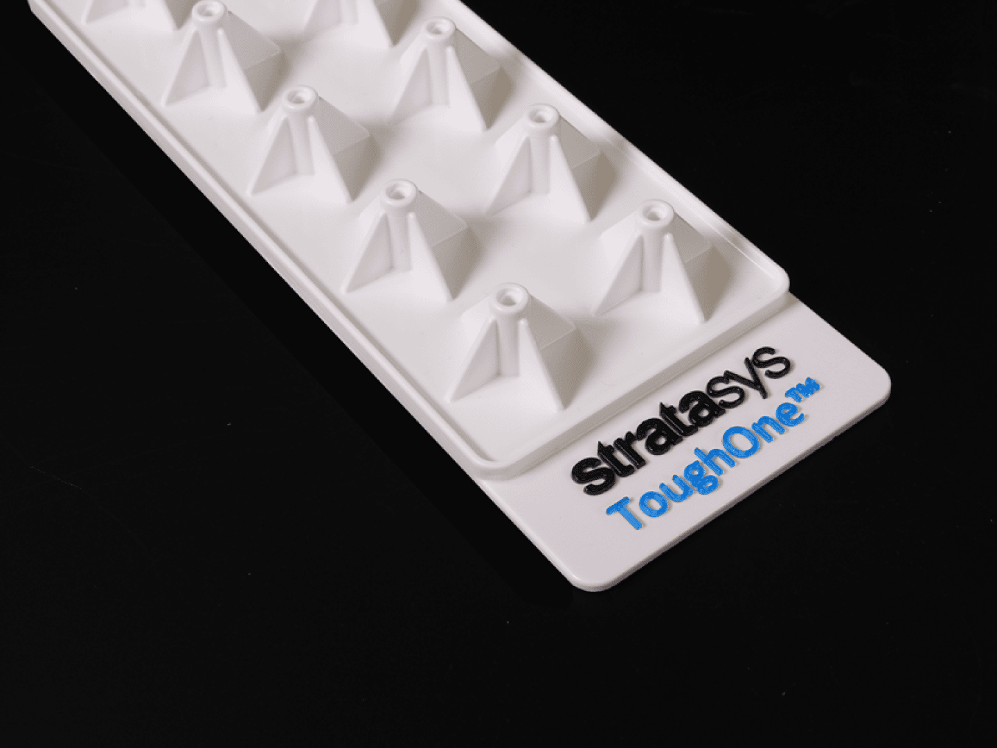 Ein weißes, rechteckiges Kunststofftablett mit pyramidenförmigen Erhebungen und dem Stratasys ToughOne™-Logo in schwarzer und blauer Schrift auf schwarzem Hintergrund.