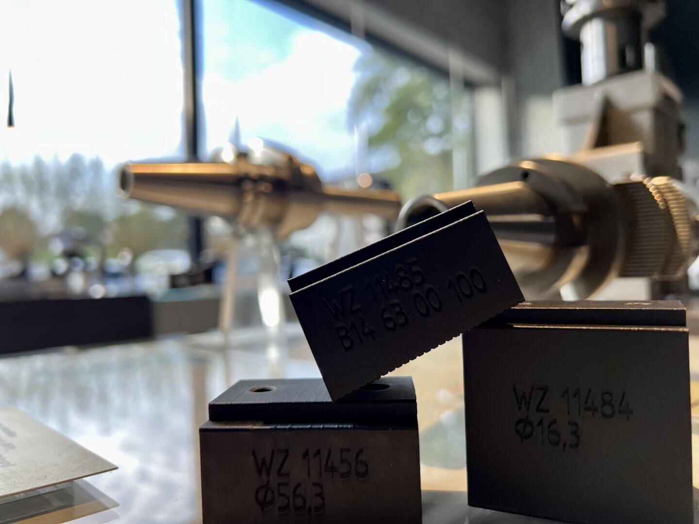 Nahaufnahme von schwarzen Metallblöcken mit eingravierten Zahlen und Buchstaben, die auf einer Glasfläche in einem hellen Raum mit unscharfen Industrieanlagen und großen Fenstern im Hintergrund ausgestellt sind.