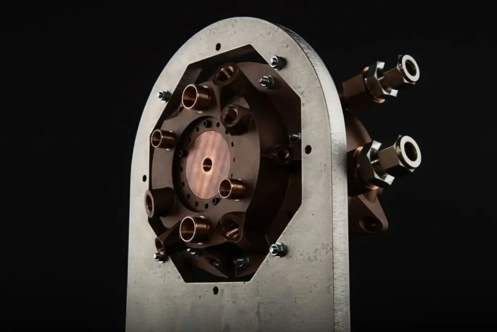 Nahaufnahme eines metallischen Industriegeräts mit mehreren Kupferdüsen und -armaturen, montiert auf einer grauen Metallplatte vor schwarzem Hintergrund.