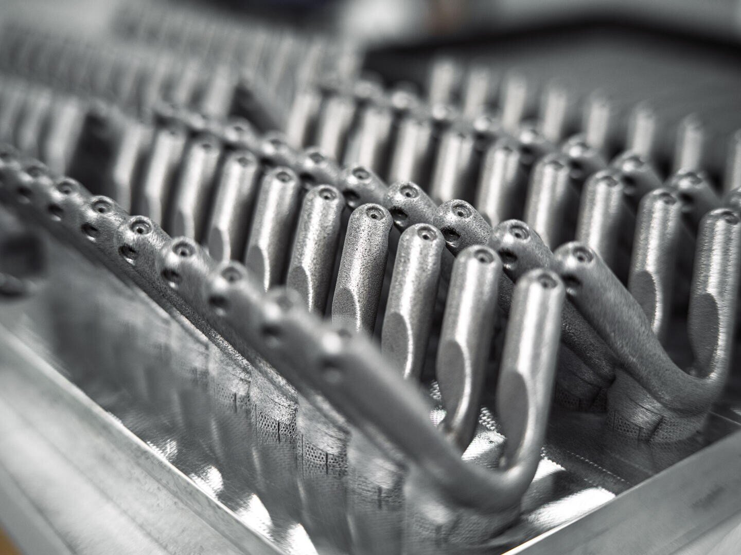 Reihen identischer, metallischer, 3D-gedruckter Teile mit abgerundeten Enden und Löchern, die in einer Industrie- oder Fertigungsumgebung sauber auf einer Metalloberfläche ausgerichtet sind.