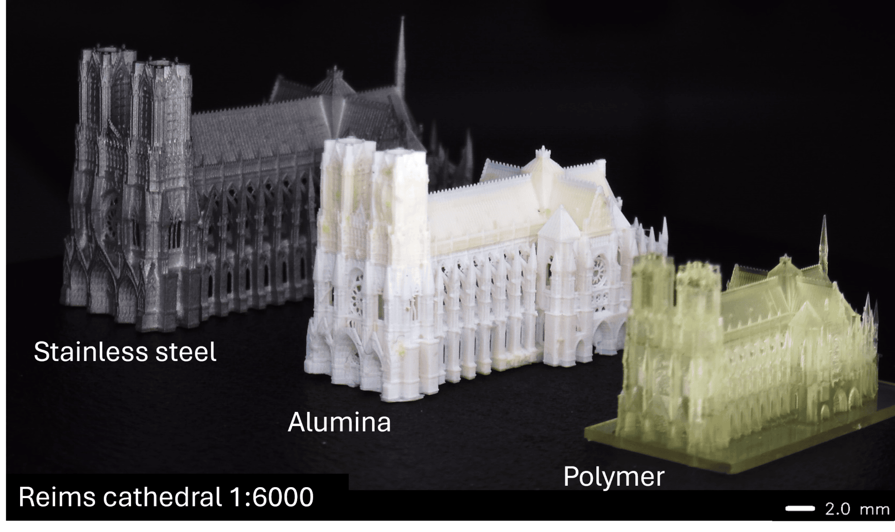 Drei detaillierte 3D-gedruckte Modelle der Kathedrale von Reims im Maßstab 1:6000 aus rostfreiem Stahl, Aluminiumoxid und Polymer werden nebeneinander auf einer schwarzen Fläche gezeigt. Es wird ein Maßstabsbalken von 2,0 mm angezeigt.