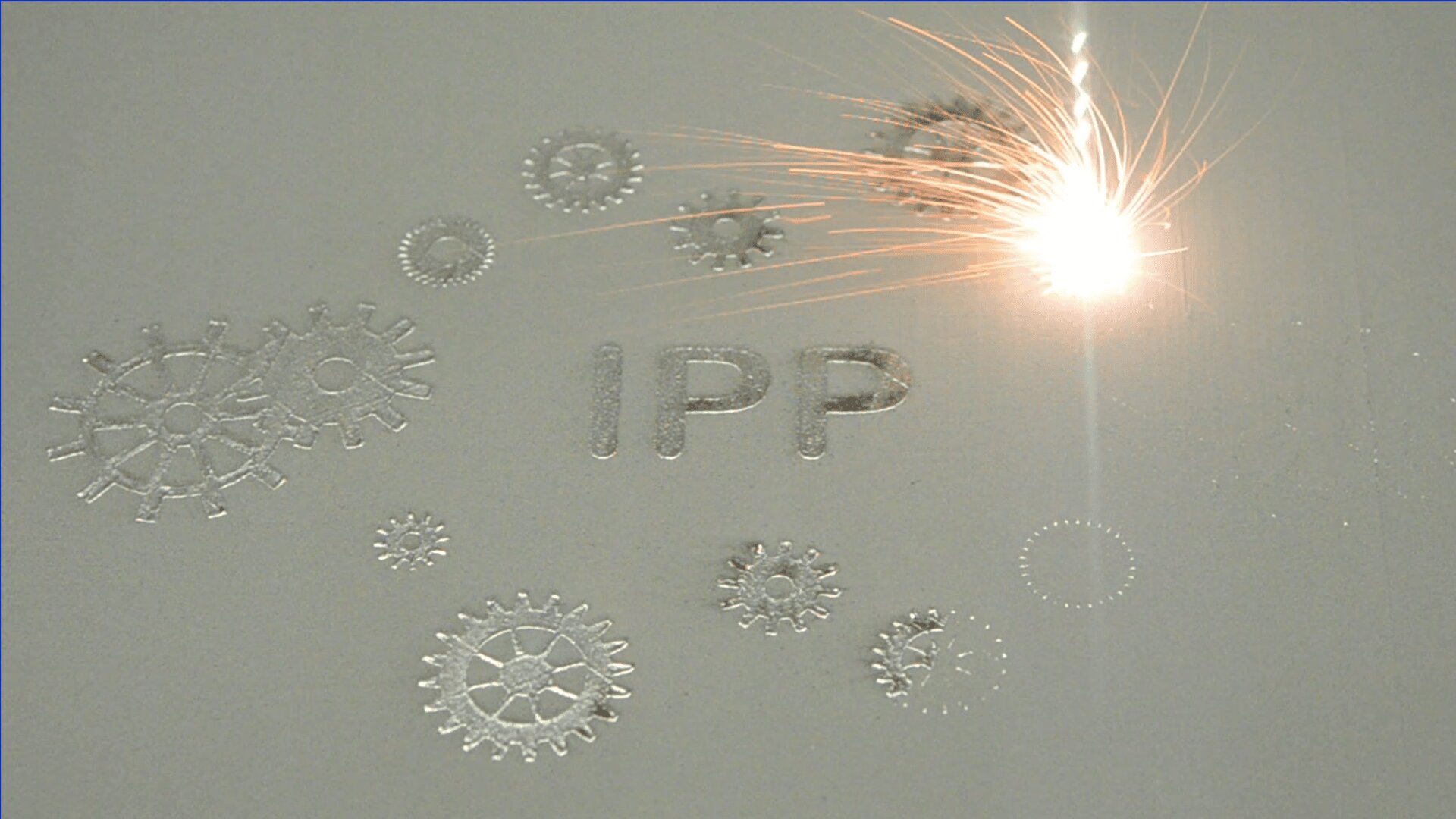 A laser beam melts heavy-duty steel in a powder bed and forms the letters "IPP" and various gear shapes, with bright sparks visible in the laser interaction zone.
