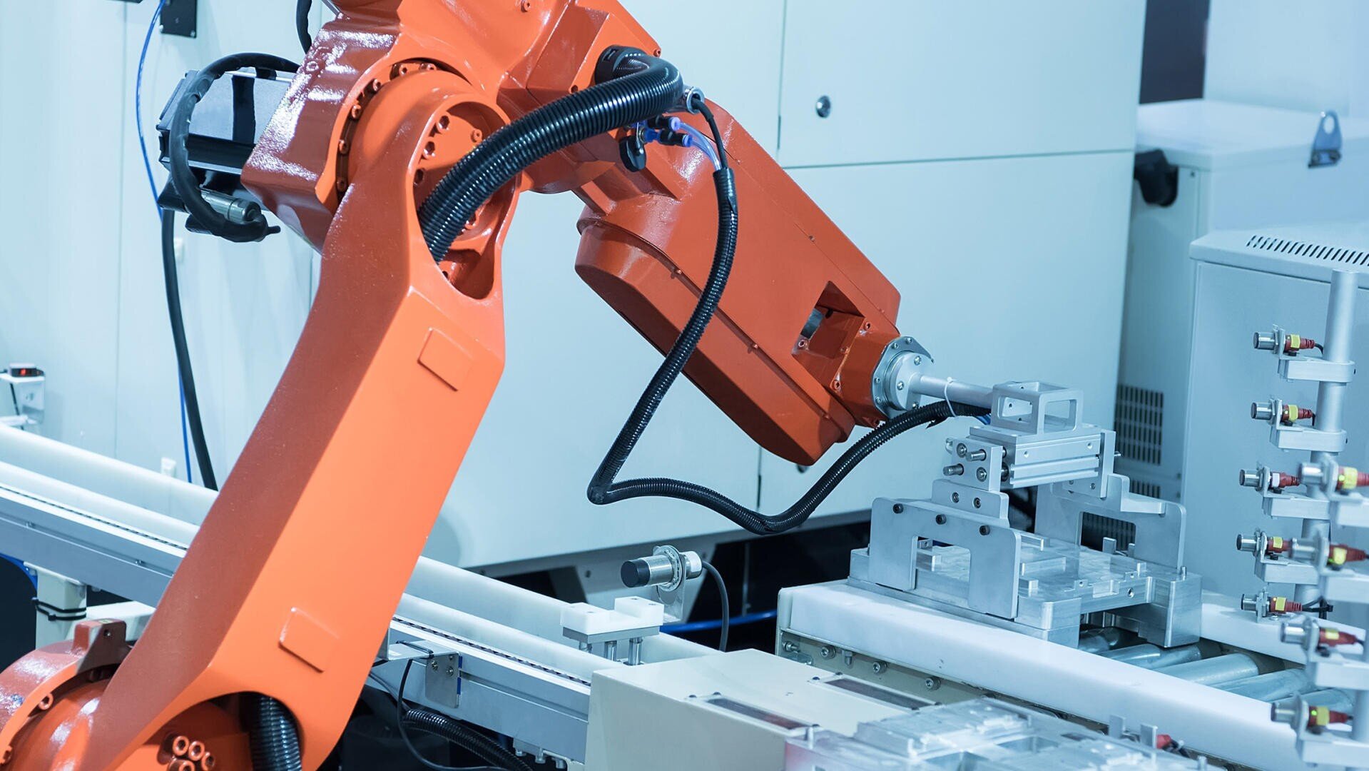 Ein orangefarbener Industrieroboterarm bedient Maschinen an einem automatisierten Fließband in einer modernen Fabrikanlage. Verschiedene Kabel und Metallteile sind zu sehen, die auf präzise Fertigungsarbeiten hinweisen.