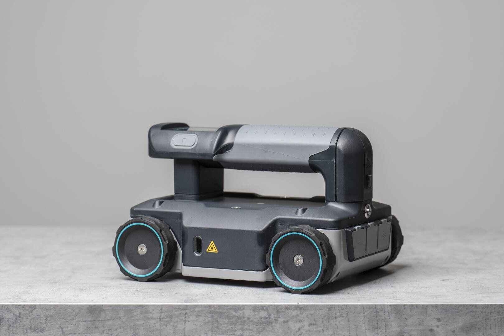 Ein kompakter Roboter mit Rädern und stabilem Griff steht auf einer grauen Fläche. Der Roboter hat vier Räder, ein dunkelgraues Gehäuse und ein Warnschild. Er scheint für industrielle oder Inspektionszwecke konzipiert zu sein.