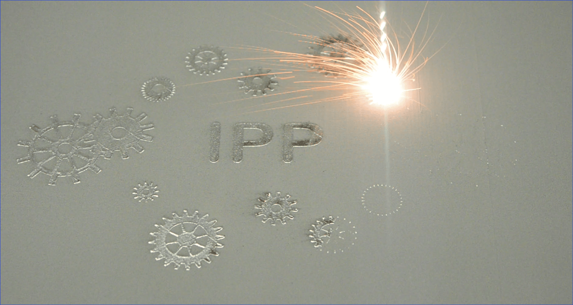 Ein Laserstrahl schmilzt hochbelastbaren Stahl in einem Pulverbett und formt die Buchstaben "IPP" und verschiedene Zahnradformen, wobei in der Laser-Wechselwirkungszone helle Funken sichtbar sind.