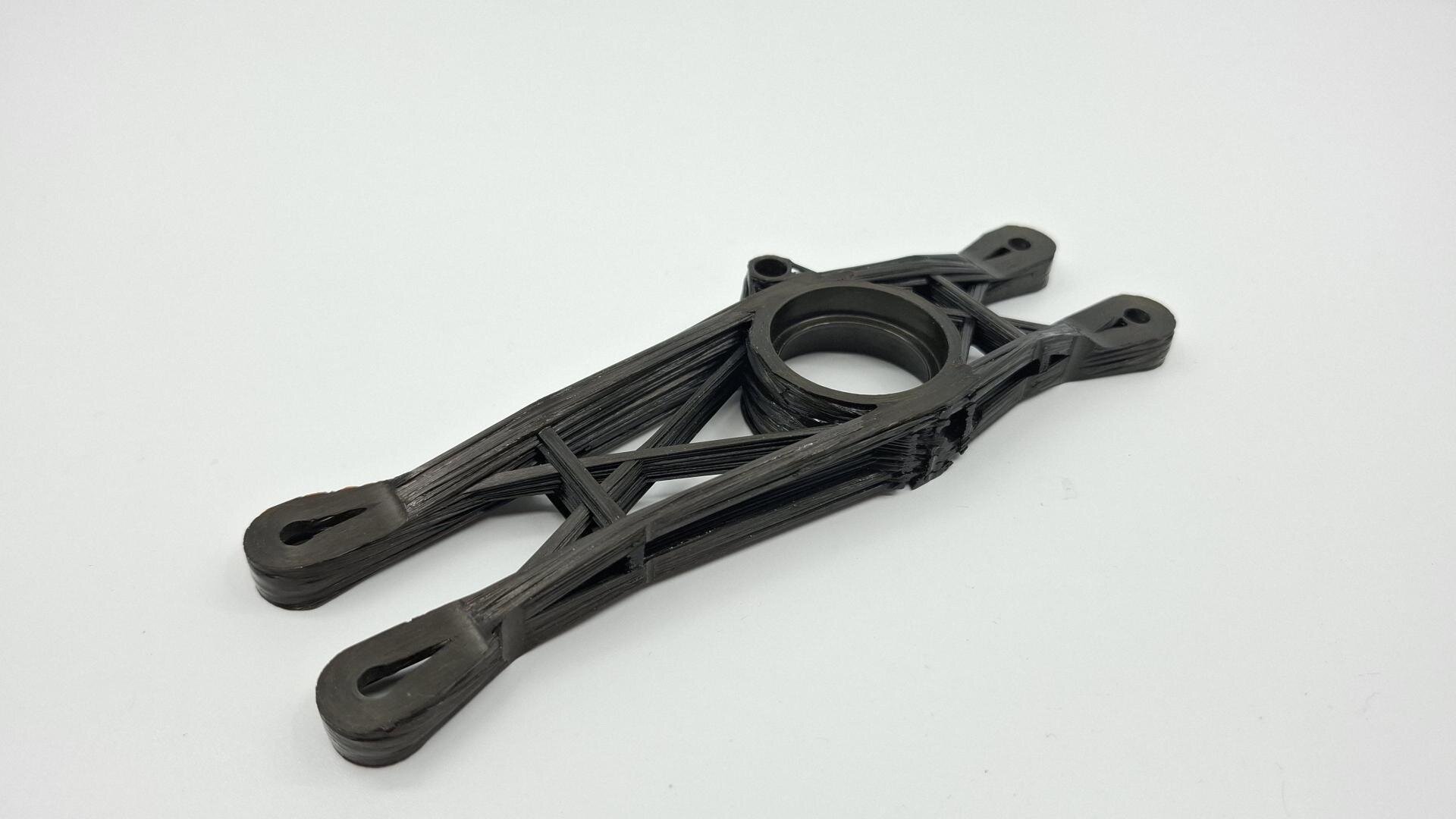 Ein schwarzes, 3D-gedrucktes Kunststoffteil mit kreisförmigem Zentrum, abgewinkelten Stützen und Löchern an jedem Ende, dargestellt auf weißem Hintergrund.