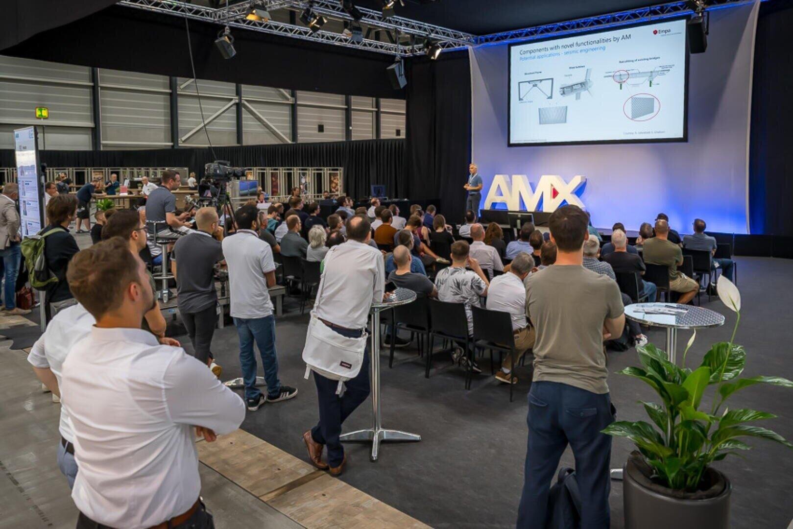 A large group of people seated and standing listen to a presentation at a conference. A presenter stands on stage near a screen displaying technical diagrams, with large illuminated AMX letters behind them.