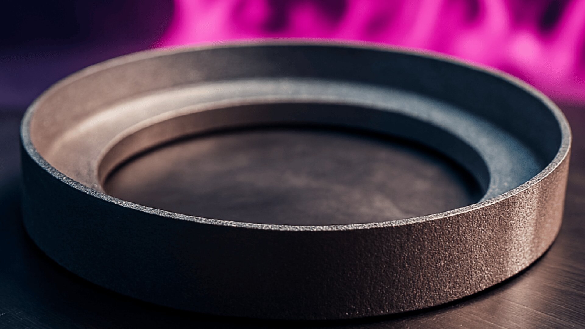 Ein runder Metallring liegt auf einer flachen, dunklen Oberfläche mit einem verschwommenen rosa und violetten Hintergrund, der an Flammen erinnert. Der Ring hat eine strukturierte, matte Oberfläche.