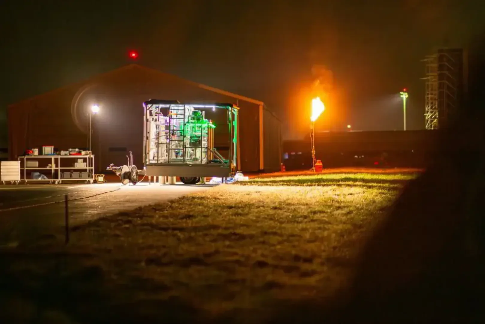Eine hell erleuchtete Industriemaschine steht nachts im Freien in der Nähe eines großen Zeltes. Im Hintergrund brennt eine große Flamme aus einem Rohr, und in der Nähe sind verschiedene Geräte verstreut.