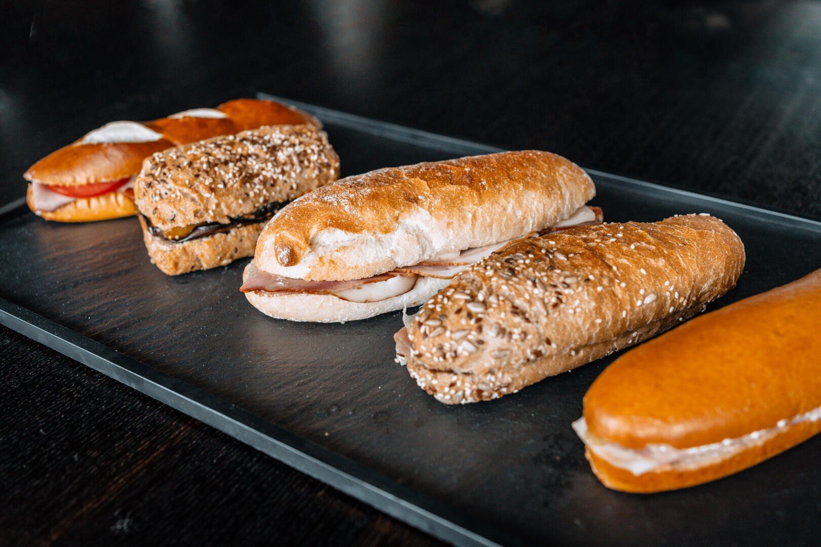 Auf einem dunklen Tablett sind verschiedene Sandwiches mit unterschiedlichen Broten und Füllungen vor einem dunklen Hintergrund aufgereiht.