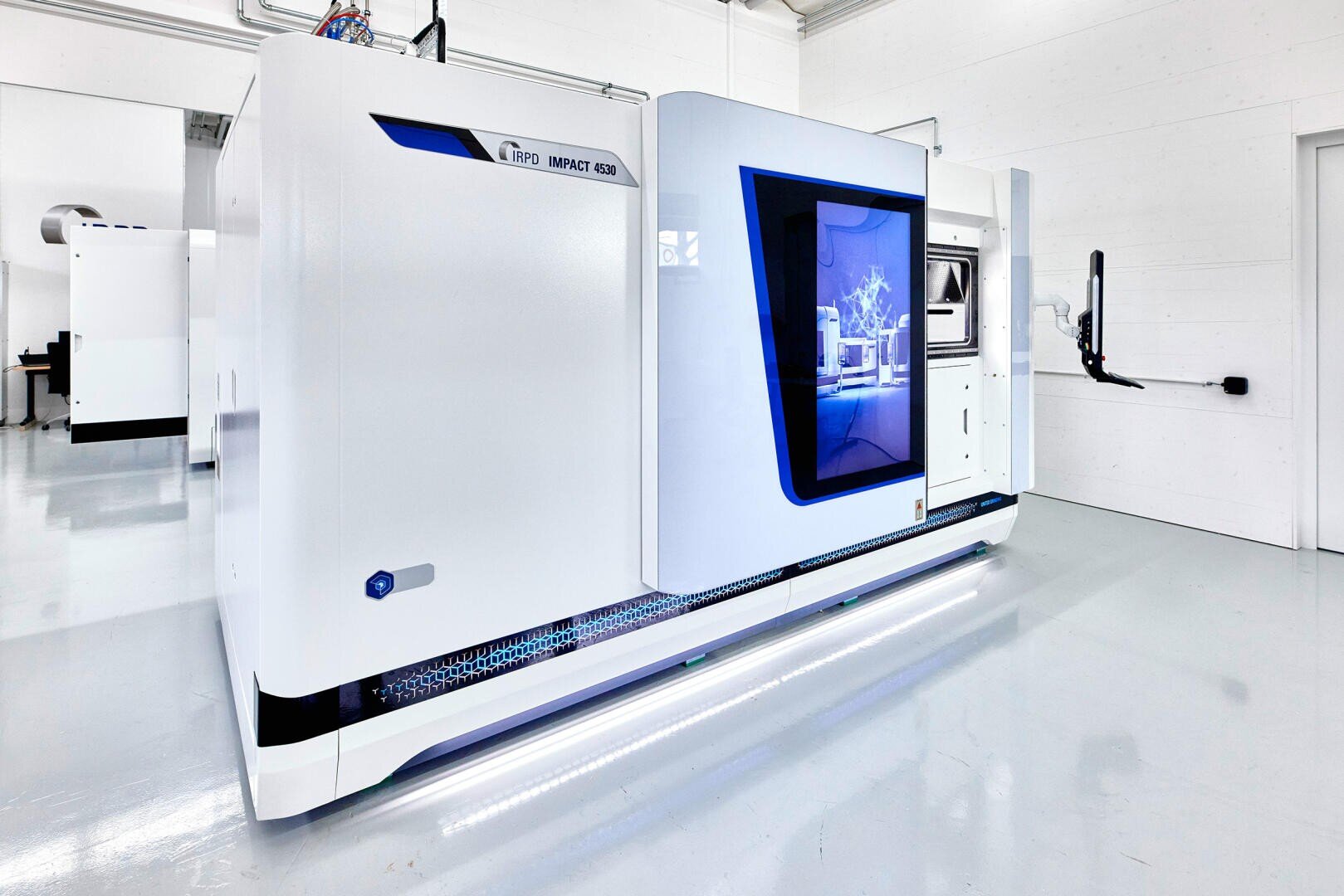 A large, modern 3D industrial printer with an elegant white and blue exterior stands in a clean, bright room with shiny floors and white walls. A screen displays a digital graphic on the side of the machine.