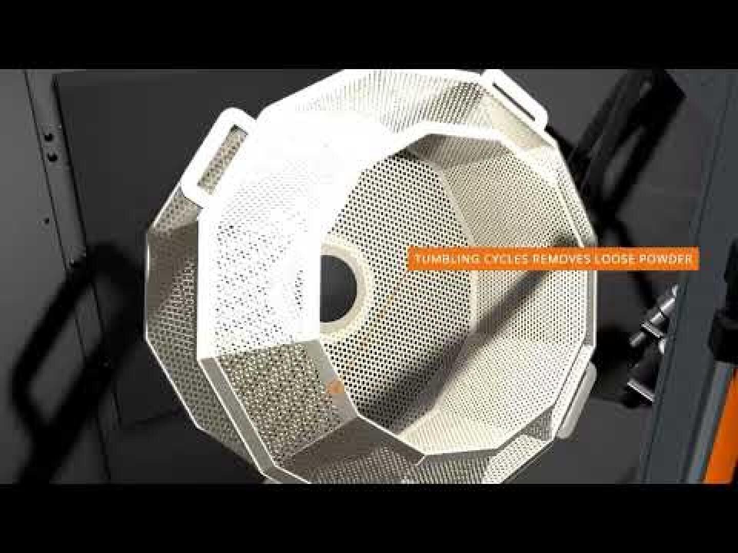 In einer Maschine rotiert eine große, weiße, perforierte Metalltrommel. Auf dem Bildschirm erscheint der Text „TUMBLING CYCLES REMOVES LOOSE POWDER“.