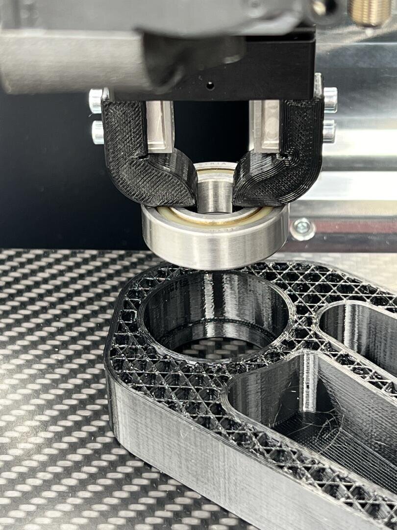 Close-up of a 3D printer head inserting a metal bearing into a partially printed black plastic part on a textured build plate.