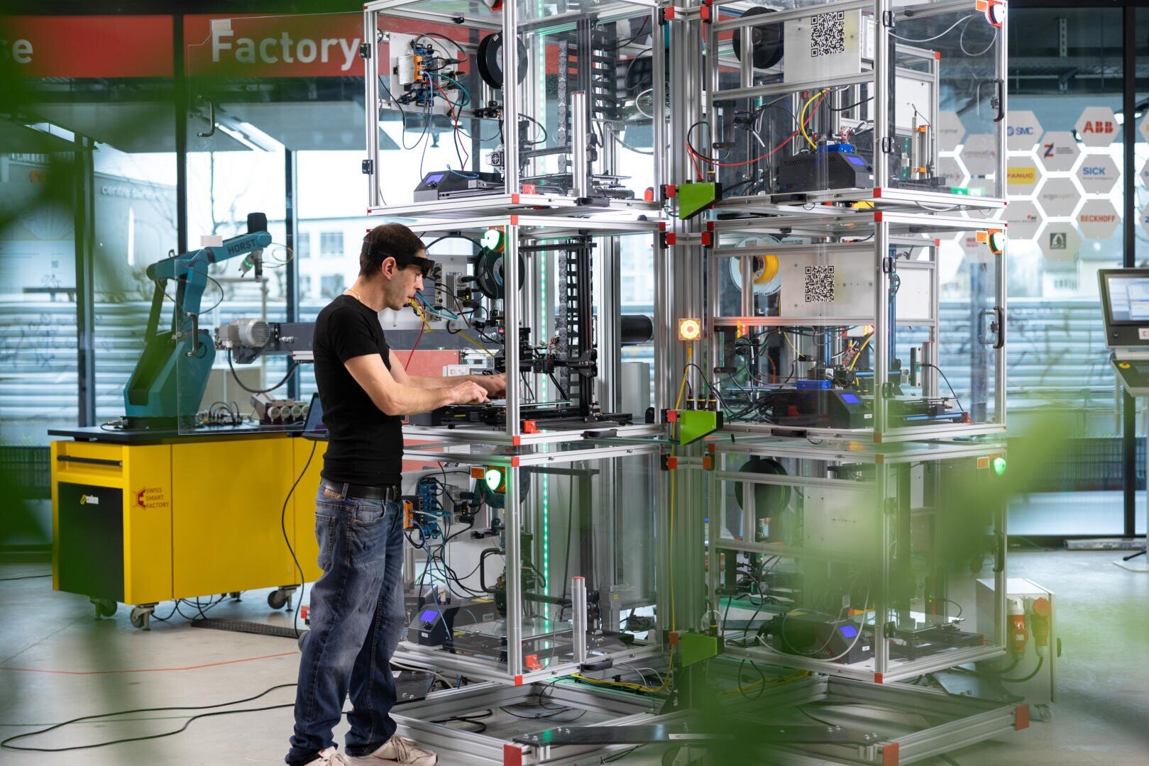 Ein Mann in schwarzem Hemd und Jeans bedient eine große, komplexe Maschine aus Metallrahmen und Drähten in einem Hightech-Labor mit Industrierobotern und Geräten im Hintergrund.