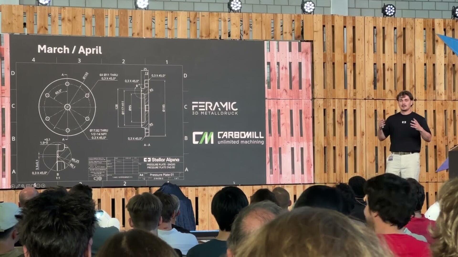 Ein Redner hält vor einem Publikum einen Vortrag vor einer großen Leinwand, auf der technische Zeichnungen und Logos von „Ceramic 3D Metalldruck“ und „Carbomill“ zu sehen sind. Im Hintergrund ist eine Wand aus Holzpaneelen zu sehen.