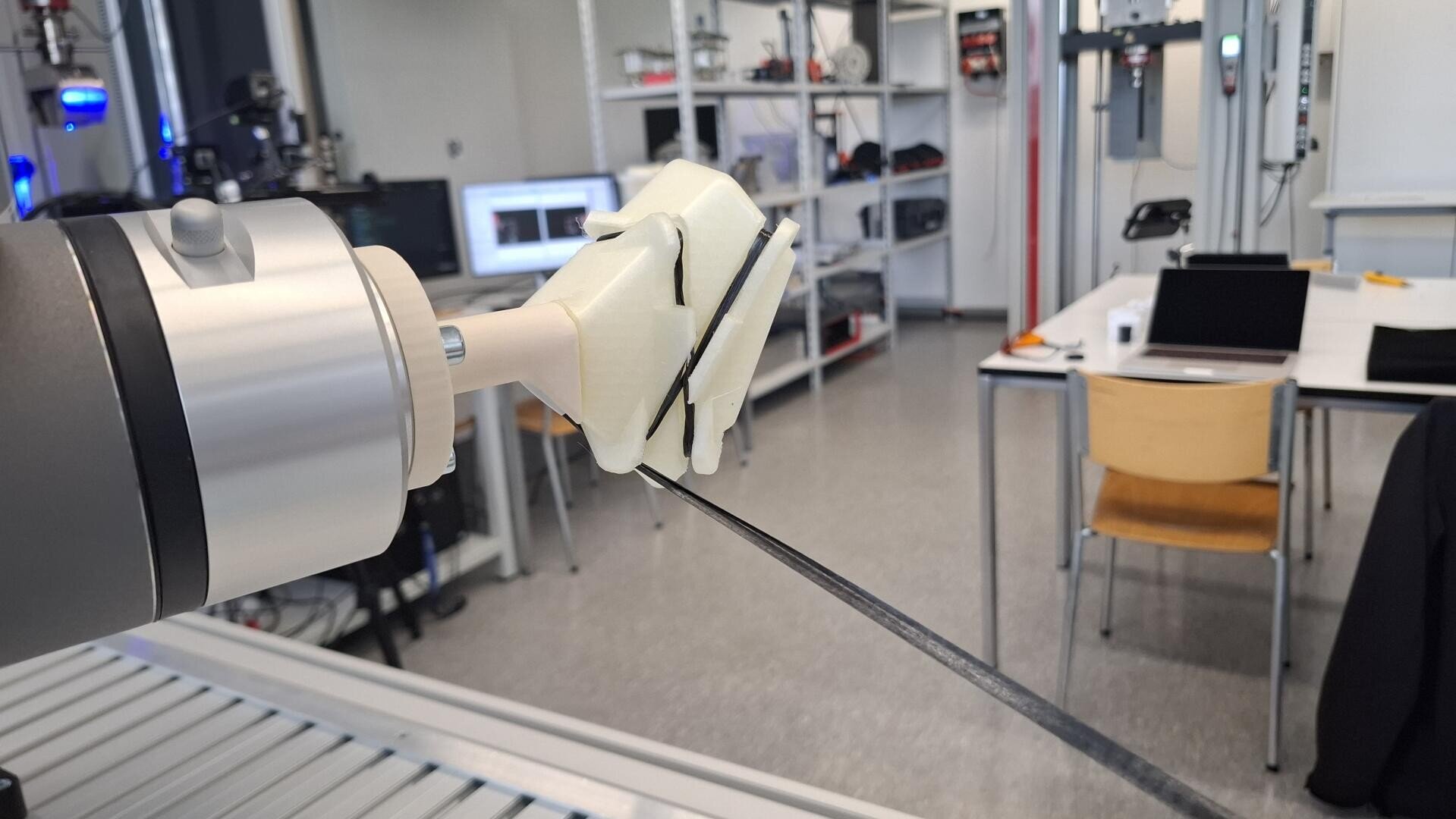 Ein Roboterarm mit einem weißen 3D-gedruckten Greifer hält einen schwarzen Streifen in einer Laborumgebung, in der im Hintergrund Tische, Stühle, Regale und Computergeräte zu sehen sind.
