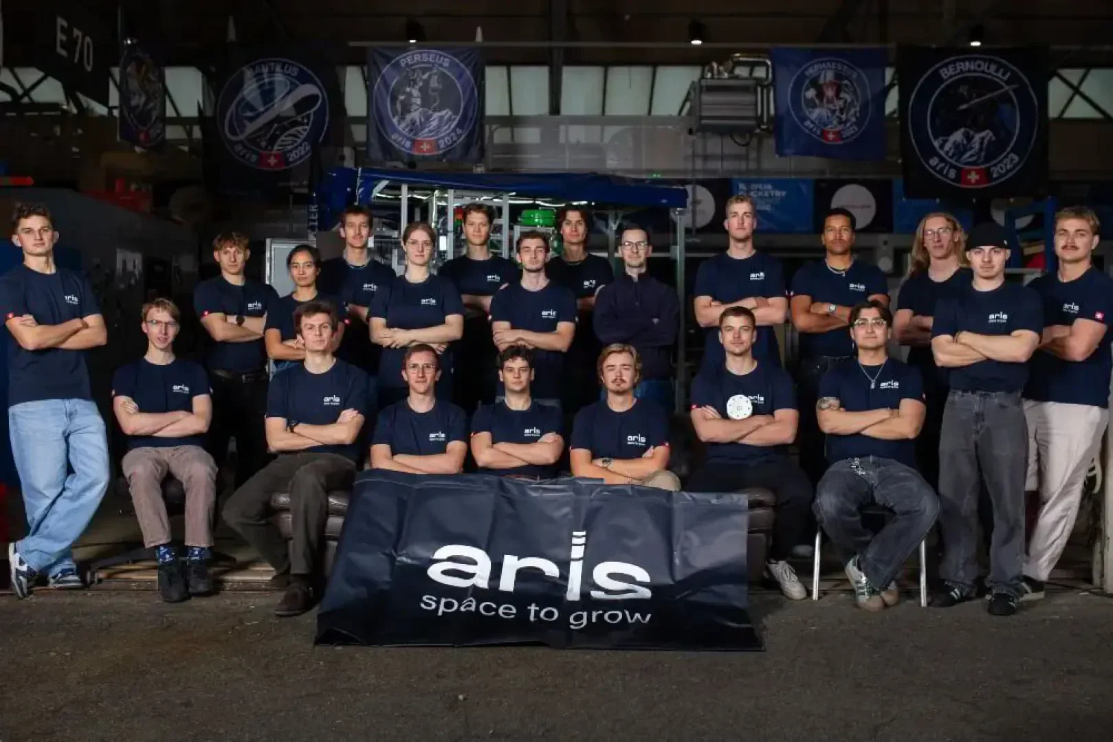 Eine Gruppe von 20 jungen Erwachsenen posiert in einem Innenraum, alle tragen passende marinefarbene ARIS-T-Shirts und stehen oder sitzen hinter einem Banner mit der Aufschrift aris space to grow. Im Hintergrund sind technische Geräte und Transparente zu sehen.